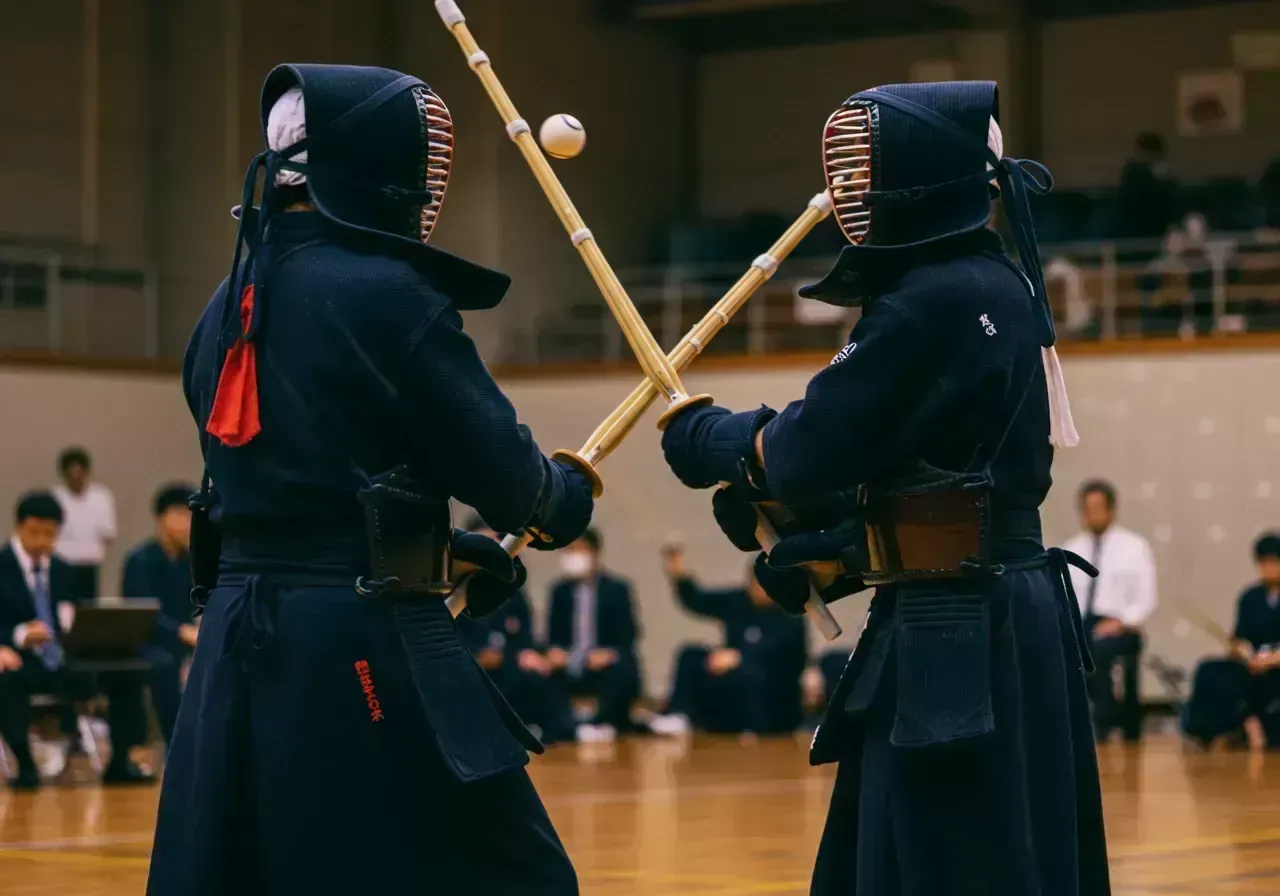 Na obrázku jsou dva cvičenci v plné kendo zbroji, kteří proti sobě zápasí s bambusovými meči v jasně osvětlené místnosti.